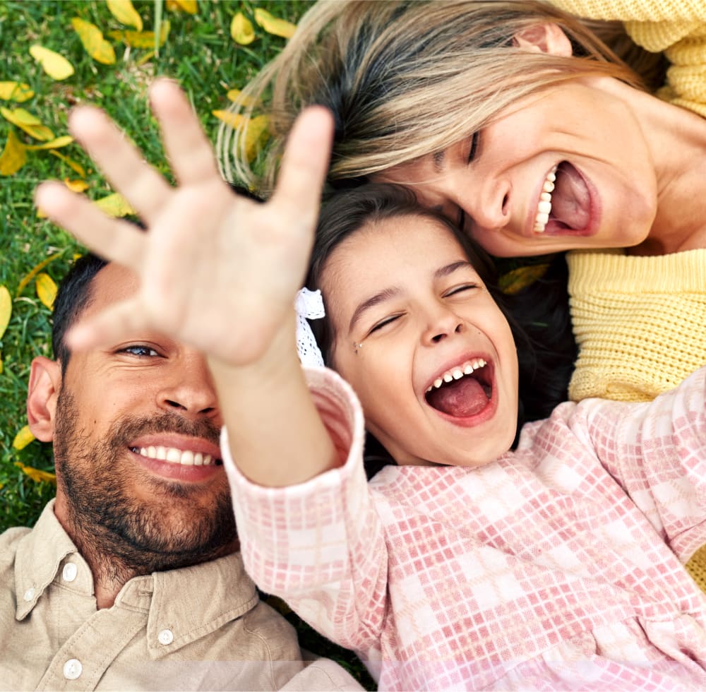 A woman with long curly hair embraces her husband who has their young daughter on his back as they all smile at the camera.