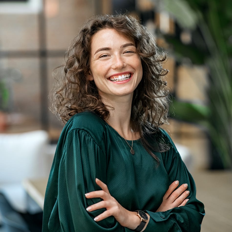 A woman with curly hair stands with her arms crossed while smiling and looking at the camera.