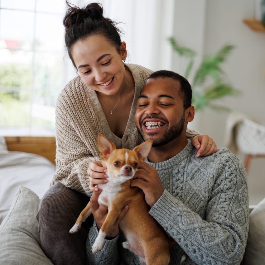 A man sits on a sofa holding a small dog and smiling while a woman stands behind him and leans over him with her arm on his shoulder.