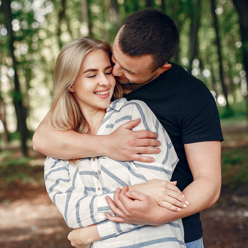 A man stands behind a woman with long hair and braces and he has his arms wrapped around her.