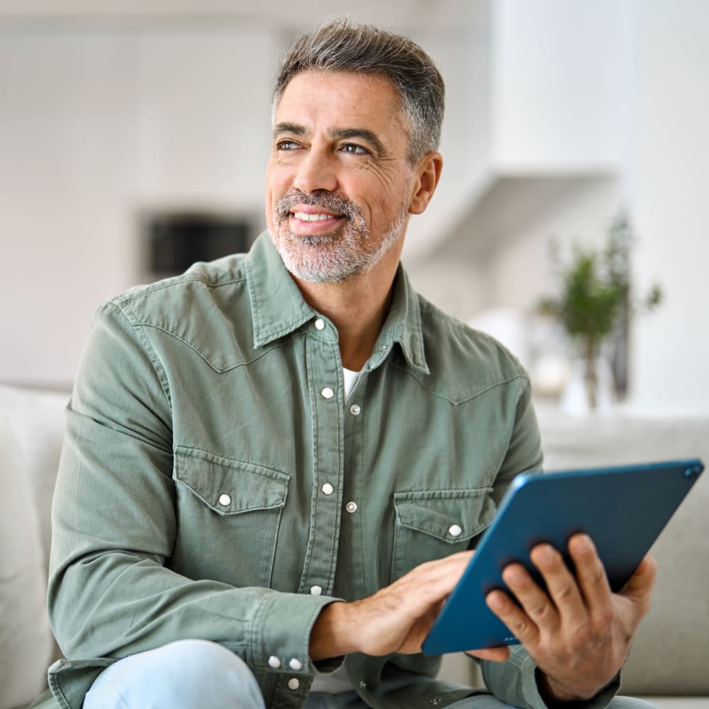 An older man sits on a sofa while holding a tablet and looking off to the side while smiling.