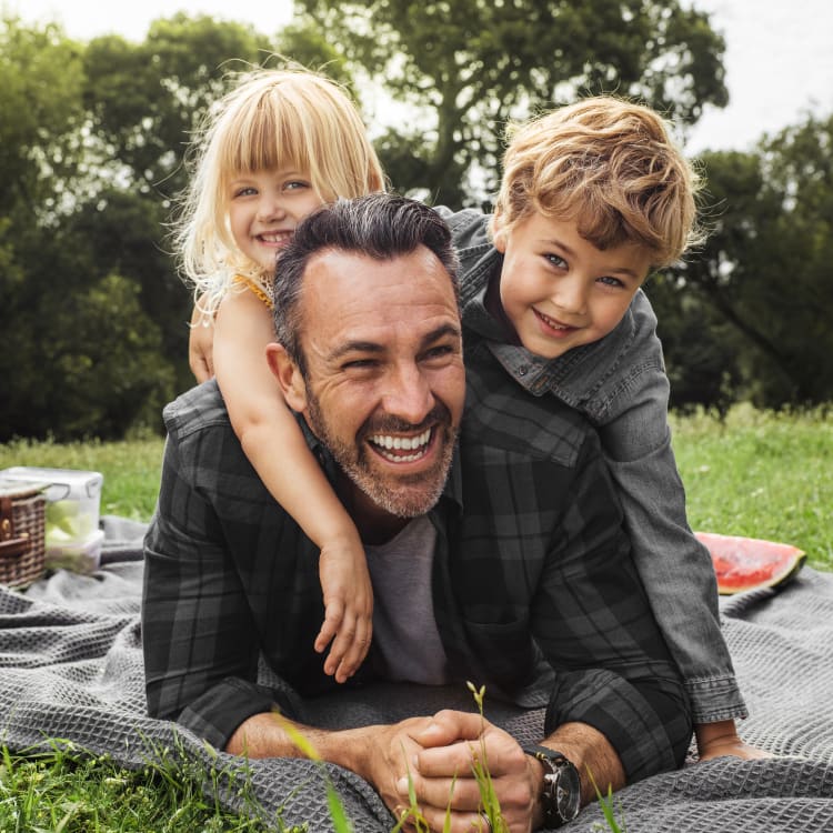 A man and two children sitting outside having a picnic. The two children and climbing onto the mans back and they are all laughing and smiling.