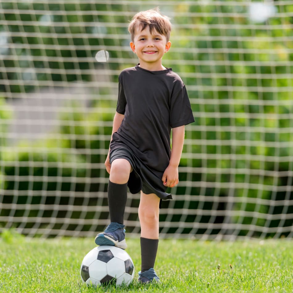 A young boy in soccer gear stands outside in front of a soccer net with his foot on top of a soccer ball.