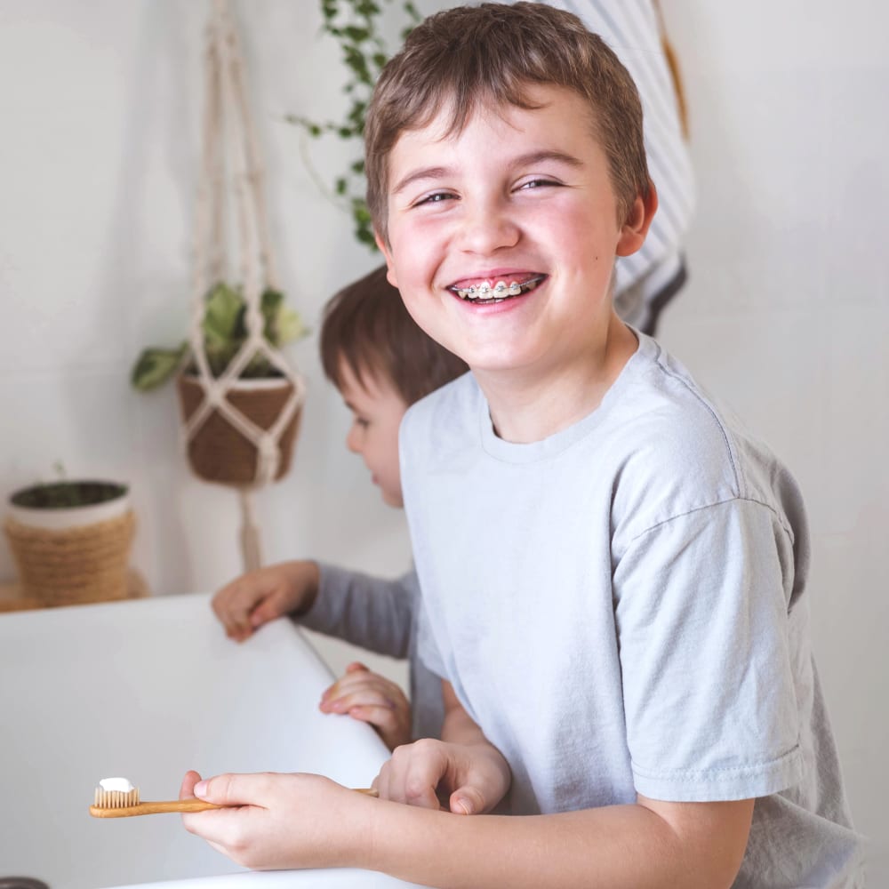 A young boy with braces stands at the bathroom sink smiling and holding a tooth brush while a younger child stands behind his just out of focus.