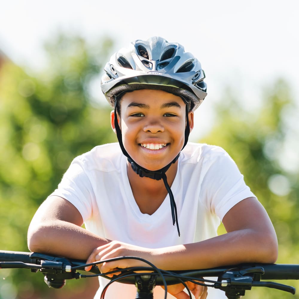 A young boy wearing a helmet stands leaning over the handles bars of his bicycle while smiling.
