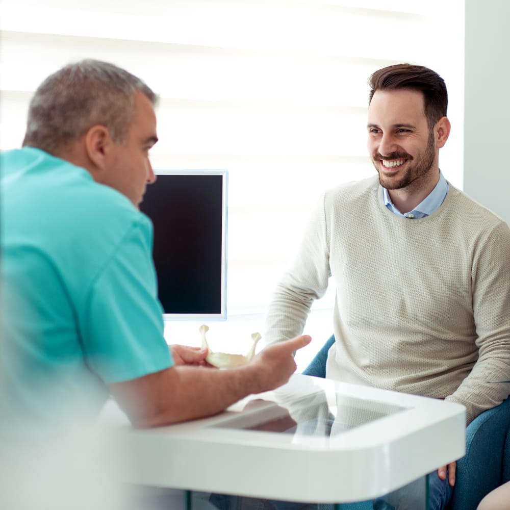 A dental professionals sits at a desk holding a model of the jaw. A male patient sits in teh chair opposite and smiles while looking at the dental professional.