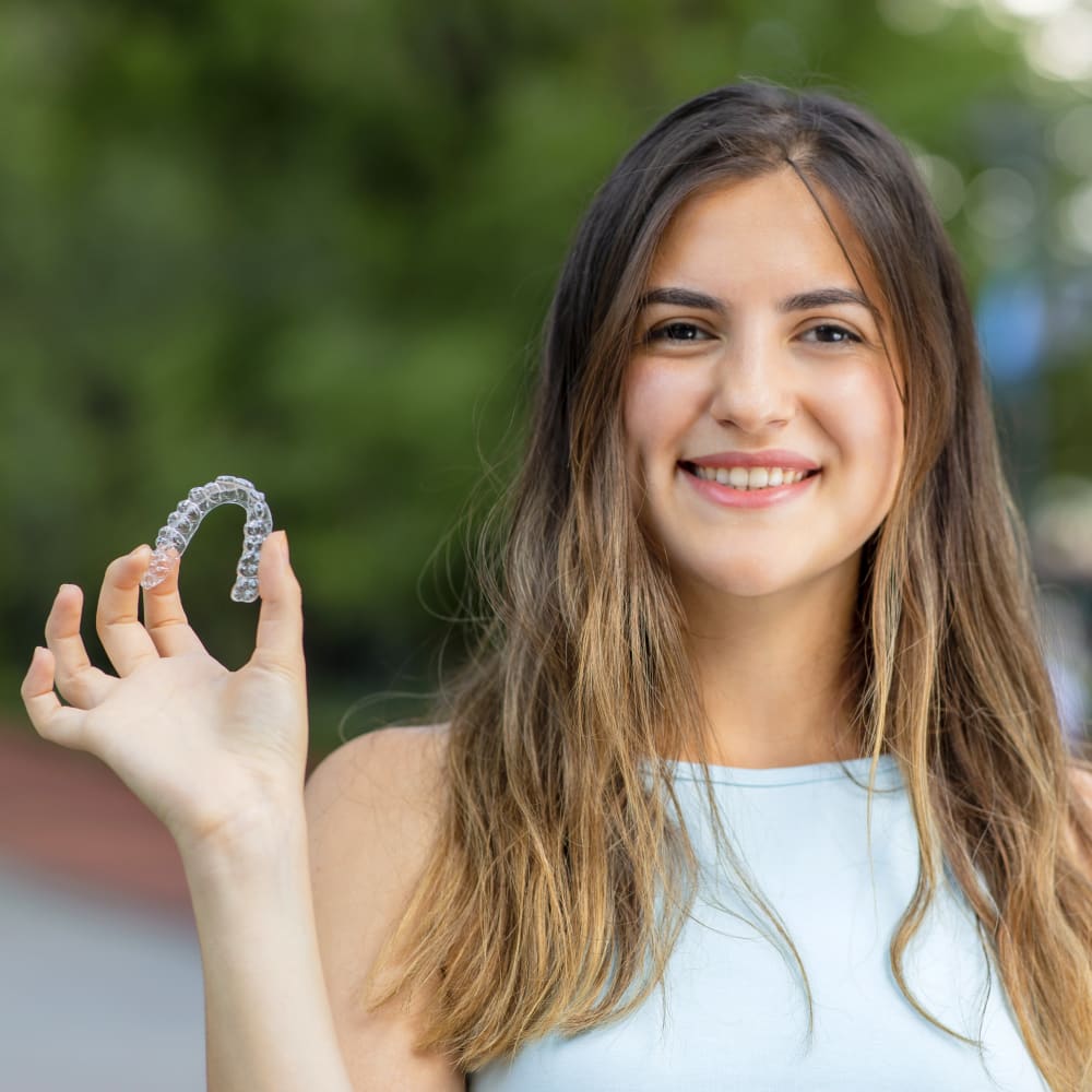 A woman stands outside in front of a treed area while smiling and holding up her Invisalign clear aligner.
