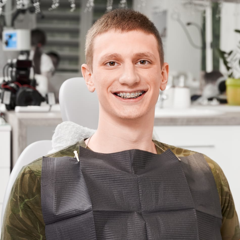 A teenage boy sits in teh chair at the clinic and smiles wide, showing off his braces.