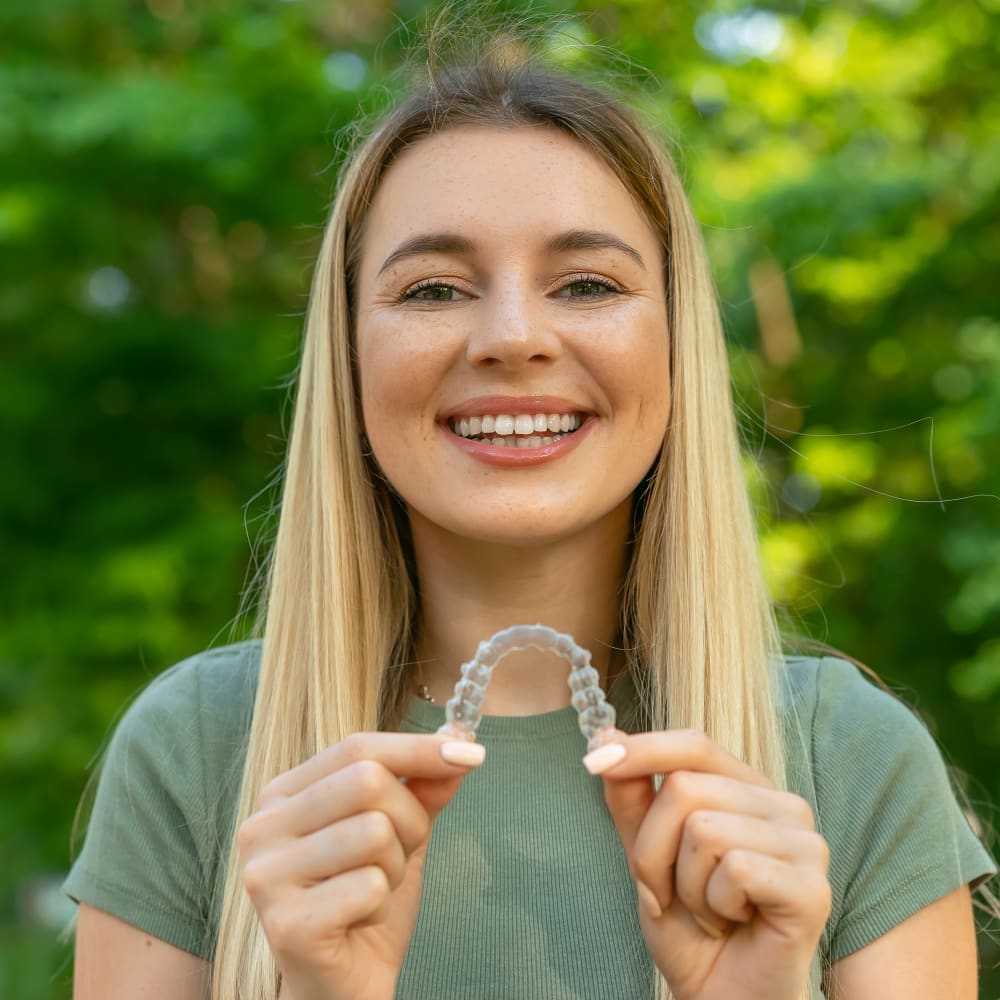 A woman is standing outside in front of green trees, smiling and holding an Invisalign clear aligner up in front of her.