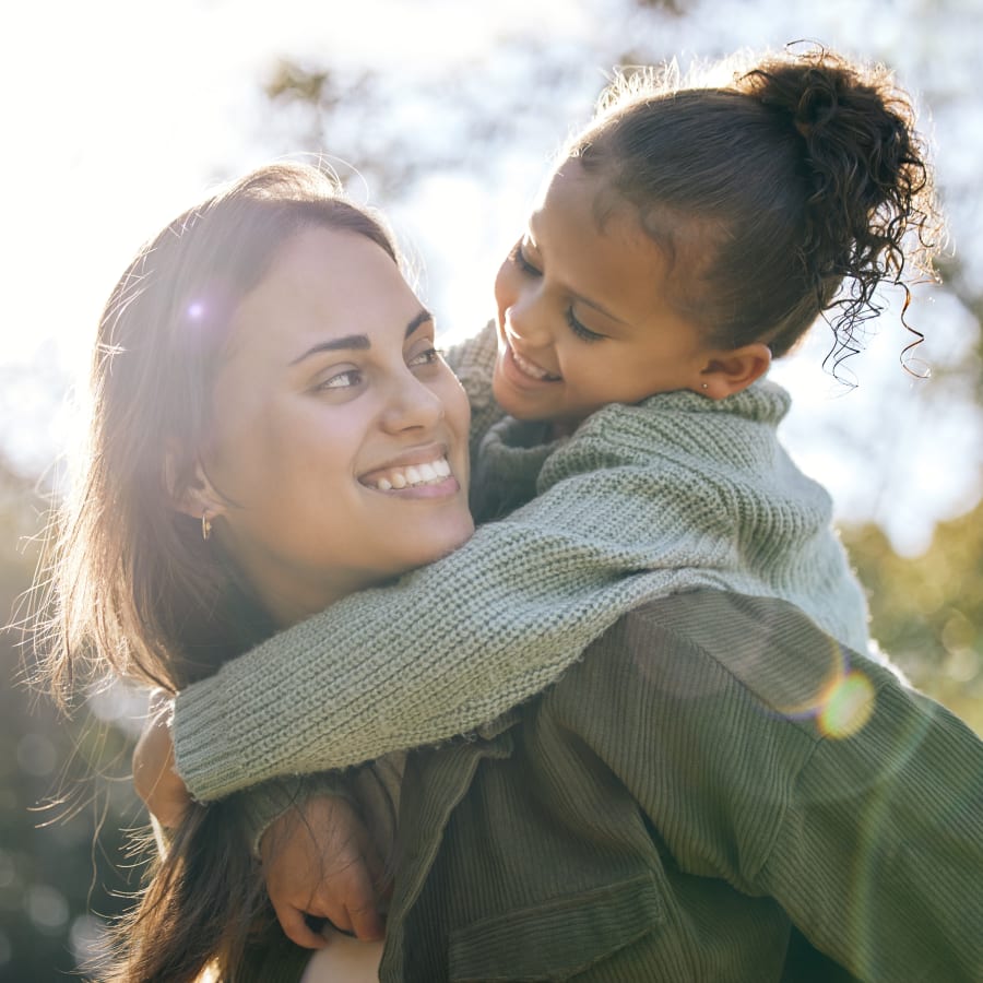 A woman stands outside while carrying a little girl on her back. The sun is shining very bright and they are smiling while lookign at eachother.