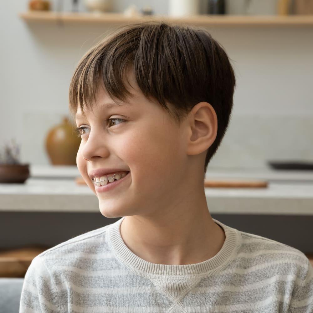 A young boy with an orthodontic retainer is looking off to the side smiling while sitting inside.