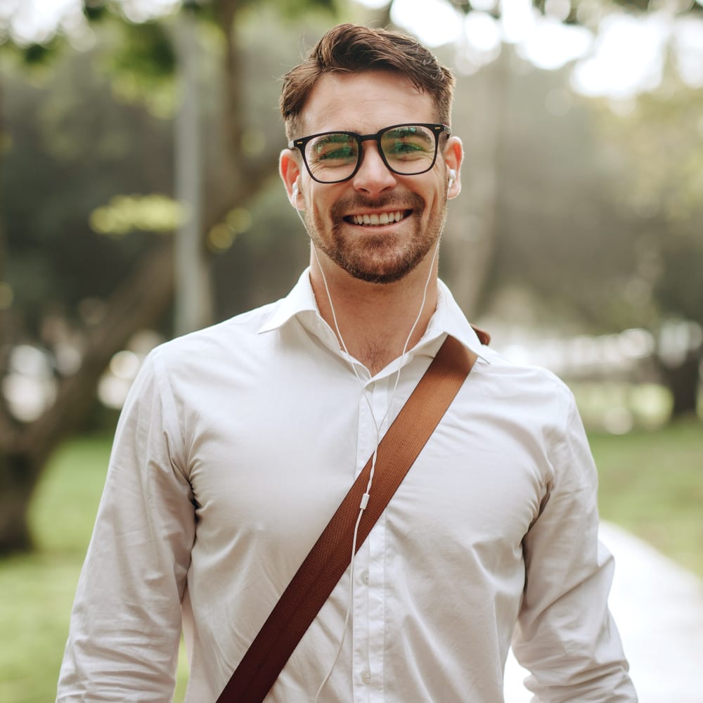 A man with glasses is standing outside with a laptop bag strap over his shoulder and headphones in while smiling.