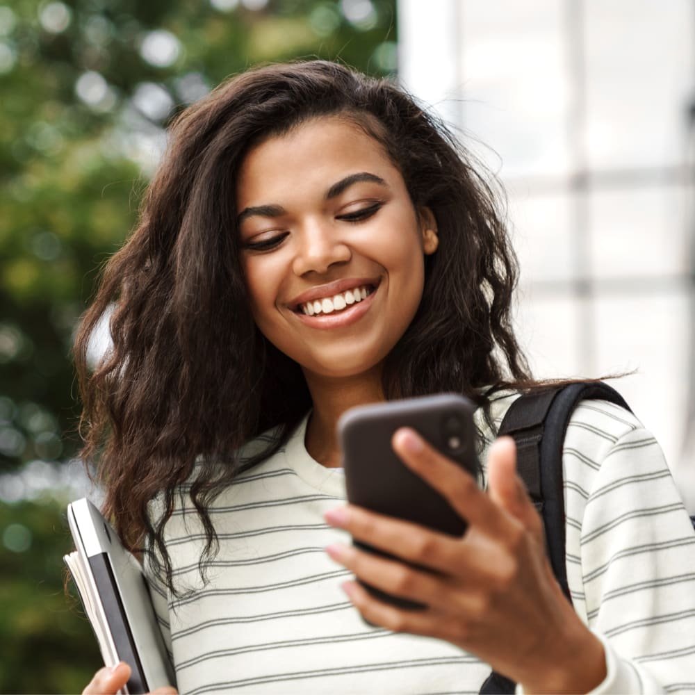 A teenage girl stands outside with a backpack over her shoulder holding books in one hand and looking down and smilng at the cellphone in her other hand.