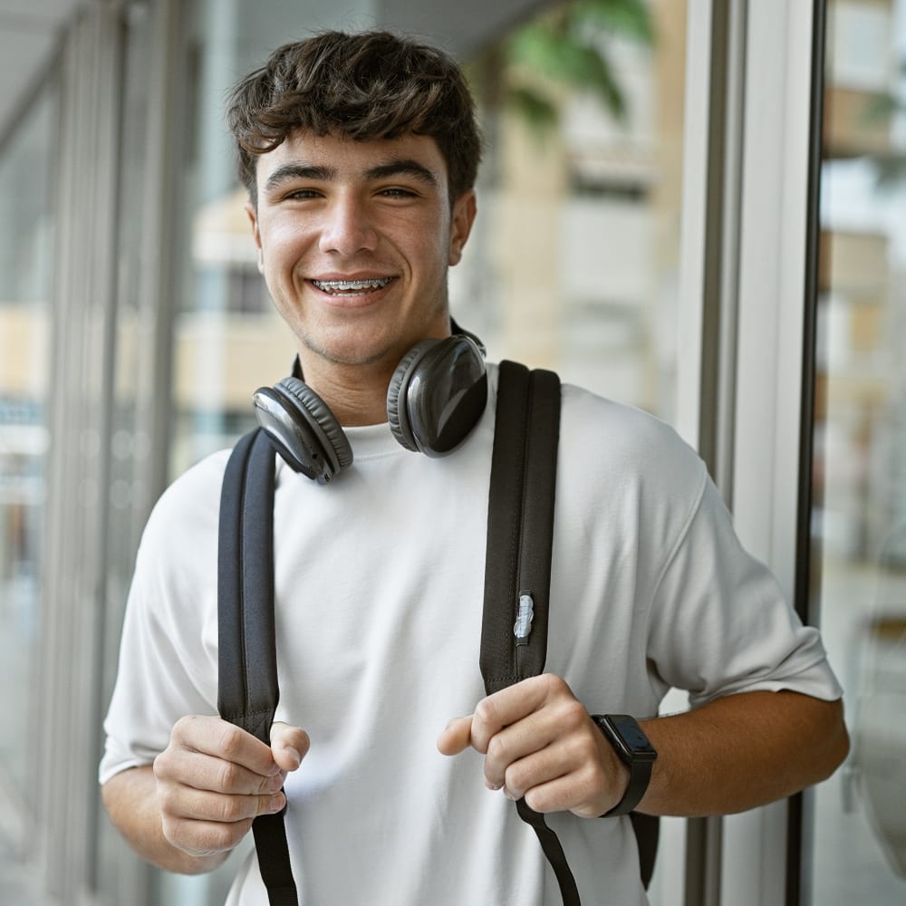 A teen boy with braces stands outside holding the straps of the backpack he is wearing. HE also has headphones around his neck and a very big smile.