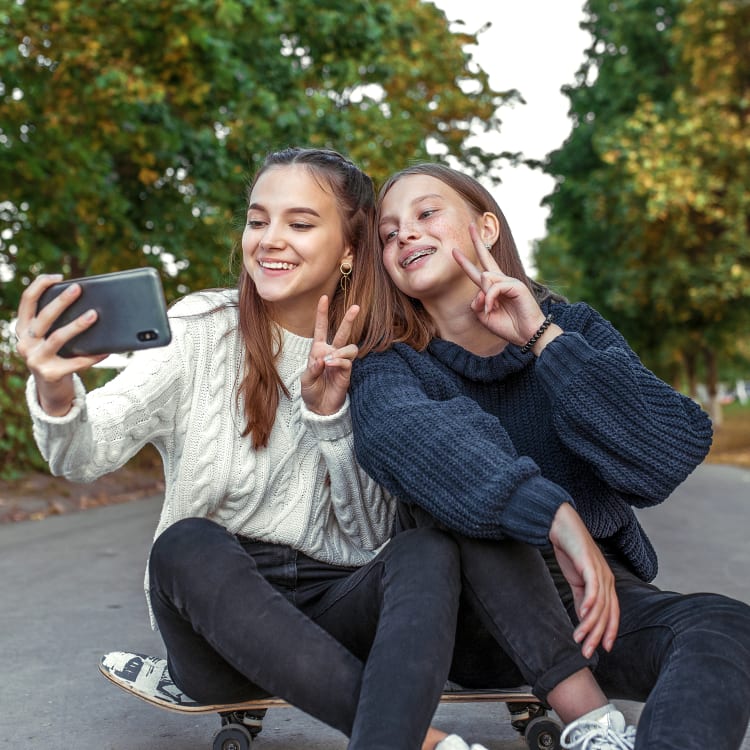 Two young teen girls sit outside on top of a skateboard and they both are smiling and holding up the peace sign while taking a selfie. One of the girls has braces.