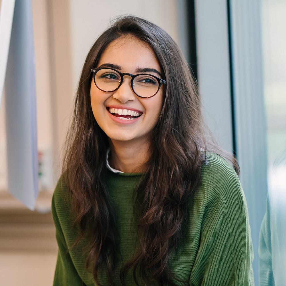 A teen girl with glasses sits inside and smiles while looking off to the side.